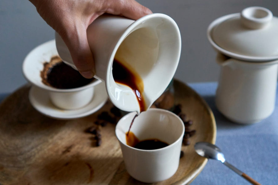 Zestaw do parzenia kawy przelewowej Slowpresso. Ręka trzyma ceramiczny dzbanek z kawą. Nalewanie kawy do czarki. Tacka drewniana.